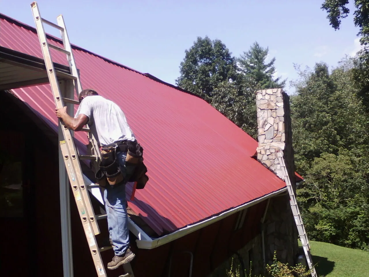 Roofer on a ladder beside a red metal roof and stone chimney — Soffit & Fascia Repair by Roofing Brothers in Fleming